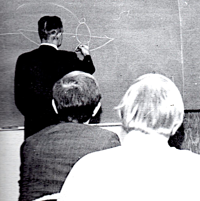 Image of brother Grazier drawing on a black board, back to camera.